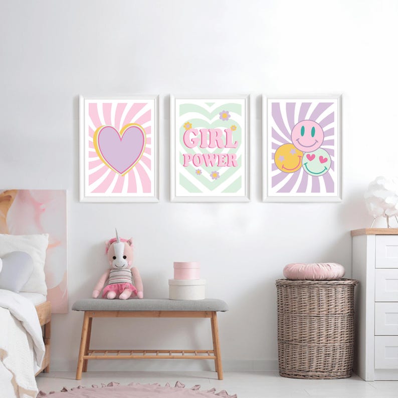 May include: Three framed wall art prints with white frames. The prints feature a heart, the words "GIRL POWER", and smiley faces. The artwork has a retro aesthetic with pastel colors and sunburst backgrounds. The prints are displayed above a bench with a stuffed unicorn.
