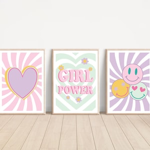 Può includere: Tre stampe artistiche incorniciate con disegni retrò. La prima presenta un cuore viola con un contorno giallo su uno sfondo a righe rosa e bianche. La seconda dice "GIRL POWER" in lettere rosa. La terza ha tre faccine sorridenti.