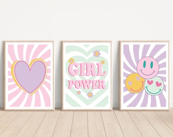 Op de afbeelding: Drie ingelijste kunstdrukken met retro-ontwerpen. De eerste heeft een paars hart met een gele omtrek tegen een roze en witte gestreepte achtergrond. De tweede zegt "GIRL POWER" in roze letters. De derde heeft drie smiley gezichten.
