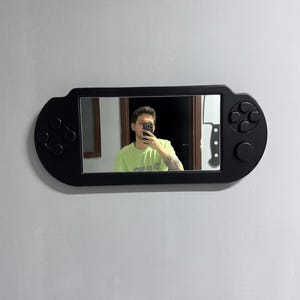 A decorative wall mirror designed in the shape of a black handheld gaming console. The frame includes raised 3D details of a D-pad, action buttons, and joysticks