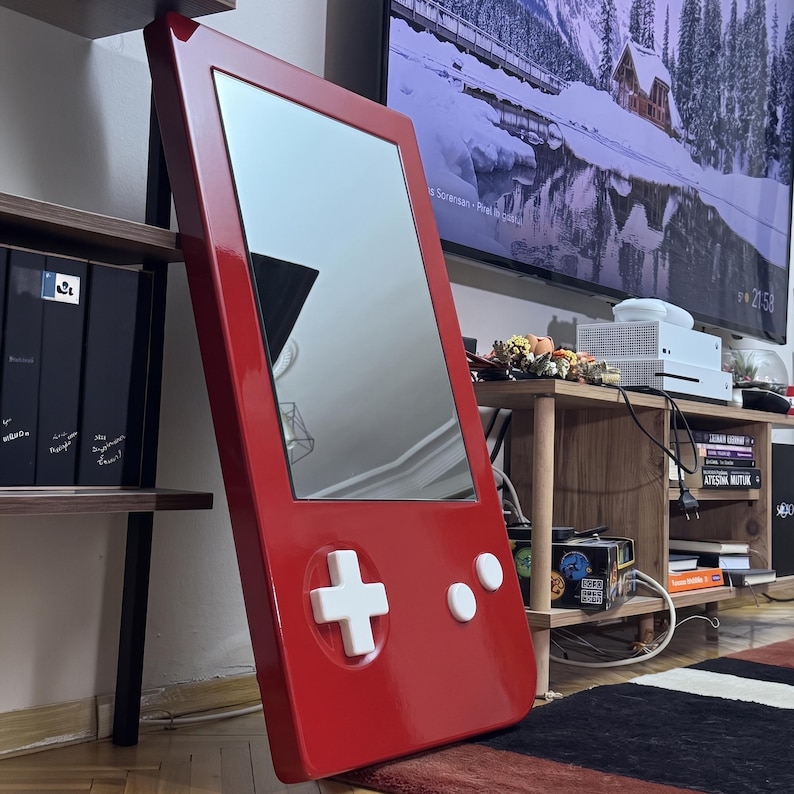 May include: A red mirror in the shape of a retro handheld video game console. The mirror features white buttons and a directional pad. The frame is glossy and leans against a wall. The mirror is approximately 30 inches tall.