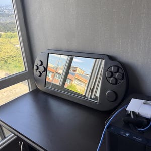 A decorative wall mirror designed in the shape of a black handheld gaming console. The frame includes raised 3D details of a D-pad, action buttons, and joysticks