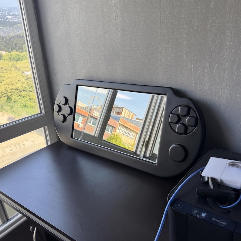 A decorative wall mirror designed in the shape of a black handheld gaming console. The frame includes raised 3D details of a D-pad, action buttons, and joysticks
