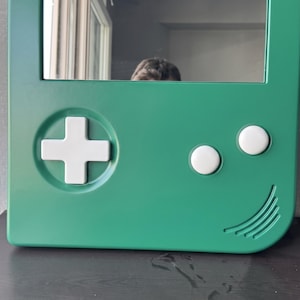 May include: Green mirror with a white cross-shaped button, two white circular buttons, and a curved line design. The mirror is shaped like a retro handheld gaming device.
