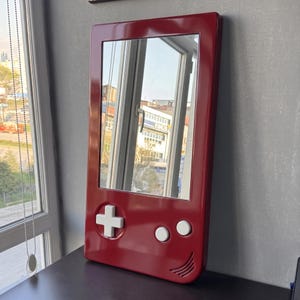 May include: A red mirror in the shape of a classic handheld video game console. The mirror has a white directional pad and two white buttons. The frame has a glossy finish and a speaker-like design at the bottom.