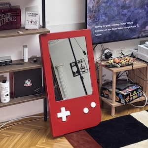 May include: A red mirror in the shape of a retro handheld game console. The mirror has white buttons and a directional pad. The mirror reflects a room with a black and white graphic on the wall.