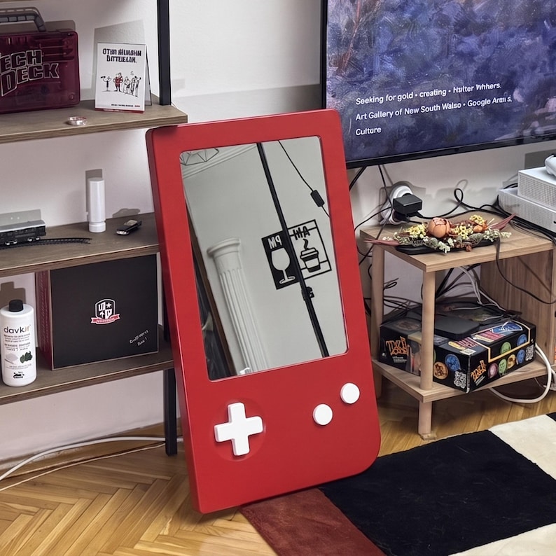 May include: A red mirror in the shape of a retro handheld game console. The mirror has white buttons and a directional pad. The mirror reflects a room with a black and white graphic on the wall.