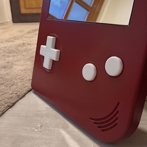 May include: A burgundy-colored mirror in the shape of a retro handheld gaming device. It features a white directional pad, two white circular buttons, and a speaker grill design. The mirror reflects a window and a room.
