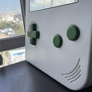 May include: A white wall-mounted mirror shaped like a classic handheld gaming device. It features a green directional pad, two green buttons, and a speaker grill design. The mirror is set against a window background.