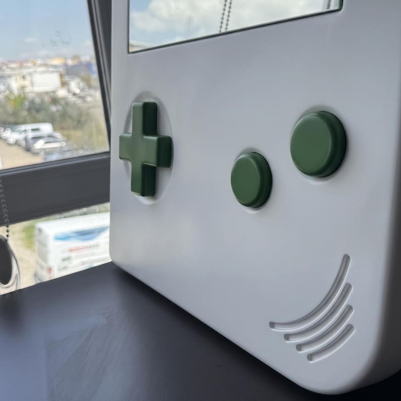 May include: A white wall-mounted mirror shaped like a classic handheld gaming device. It features a green directional pad, two green buttons, and a speaker grill design. The mirror is set against a window background.