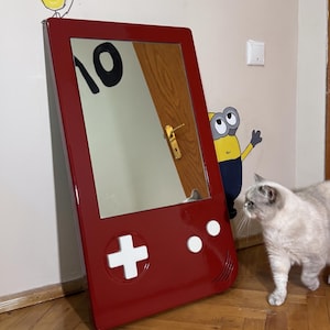 May include: A red framed mirror designed to resemble a classic handheld video game console. The mirror features white control buttons and a directional pad. The mirror is mounted on a wall, reflecting the room's interior.