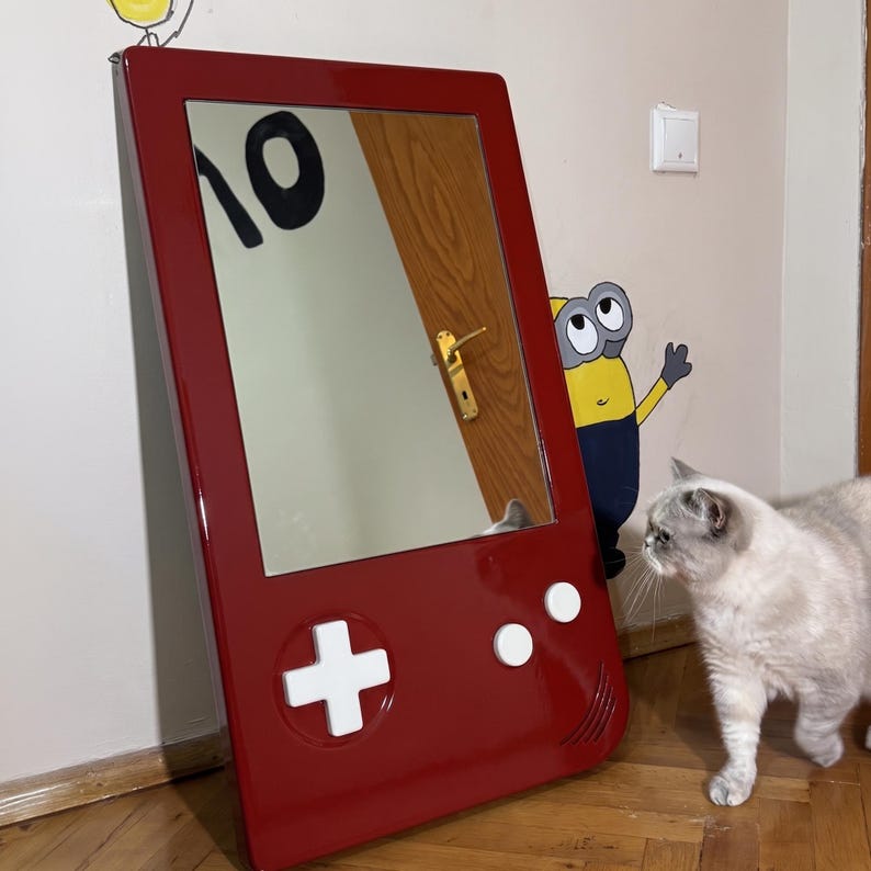 May include: A red framed mirror designed to resemble a classic handheld video game console. The mirror features white control buttons and a directional pad. The mirror is mounted on a wall, reflecting the room's interior.