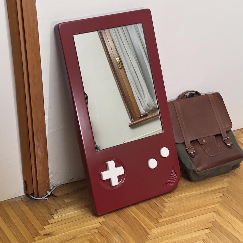 May include: A burgundy-colored mirror in the shape of a retro handheld video game console. The mirror has white buttons and a directional pad. A brown and green backpack rests on the wooden floor next to the mirror.