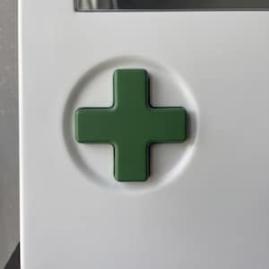 May include: A green cross emblem set in a white circular recess. The cross is a solid, dark green color, and the background is a clean, bright white. The design is simple and modern.