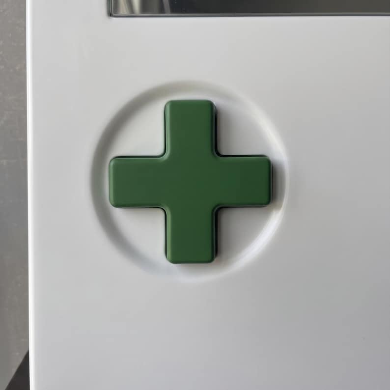 May include: A green cross emblem set in a white circular recess. The cross is a solid, dark green color, and the background is a clean, bright white. The design is simple and modern.