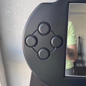 A decorative wall mirror designed in the shape of a black handheld gaming console. The frame includes raised 3D details of a D-pad, action buttons, and joysticks