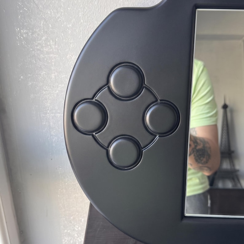 A decorative wall mirror designed in the shape of a black handheld gaming console. The frame includes raised 3D details of a D-pad, action buttons, and joysticks