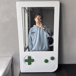 May include: White wall mirror in the shape of a classic handheld video game console. The mirror features a green directional pad and two green buttons. The mirror is rectangular with rounded corners and a curved bottom edge.