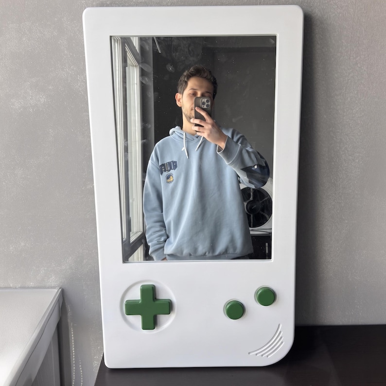 May include: White wall mirror in the shape of a classic handheld video game console. The mirror features a green directional pad and two green buttons. The mirror is rectangular with rounded corners and a curved bottom edge.