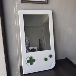 May include: A white mirror in the shape of a retro handheld gaming device. It features a green directional pad and green buttons. The mirror is rectangular with rounded edges and a reflective surface.