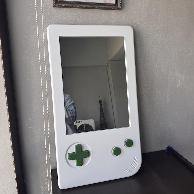 May include: A white mirror in the shape of a retro handheld gaming device. It features a green directional pad and green buttons. The mirror is rectangular with rounded edges and a reflective surface.