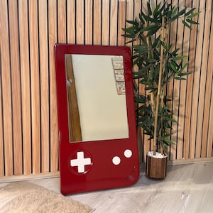 May include: A red framed mirror shaped like a retro handheld game console. The mirror has white buttons and a plus sign on the front. The mirror is leaning against a wooden wall.