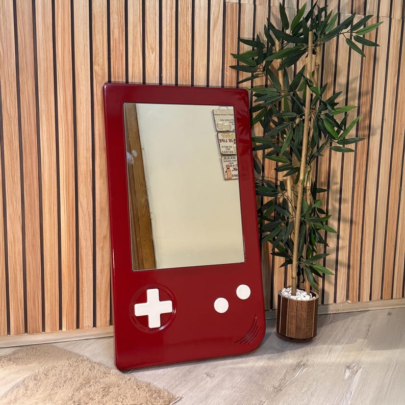 May include: A red framed mirror shaped like a retro handheld game console. The mirror has white buttons and a plus sign on the front. The mirror is leaning against a wooden wall.