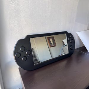 A decorative wall mirror designed in the shape of a black handheld gaming console. The frame includes raised 3D details of a D-pad, action buttons, and joysticks
