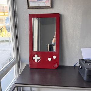 May include: A red rectangular mirror with a white cross, two white dots, and a curved line, resembling a handheld gaming device. The mirror is mounted on a gray wall above a dark brown desk.