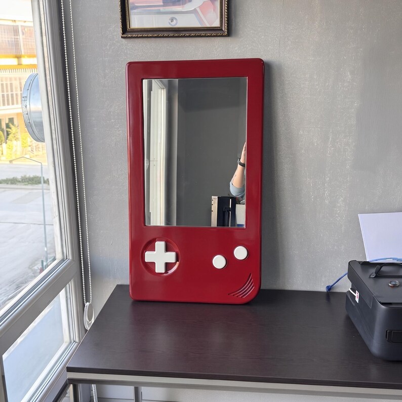 May include: A red rectangular mirror with a white cross, two white dots, and a curved line, resembling a handheld gaming device. The mirror is mounted on a gray wall above a dark brown desk.