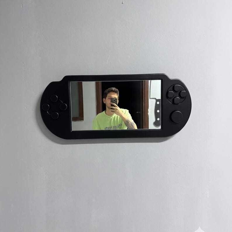 A decorative wall mirror designed in the shape of a black handheld gaming console. The frame includes raised 3D details of a D-pad, action buttons, and joysticks