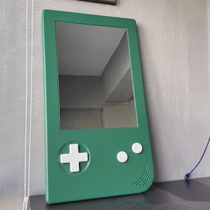 May include: A green mirror shaped like a handheld video game console. The mirror has a rectangular reflective surface and features white buttons and a directional pad. The frame is a solid, vibrant green.