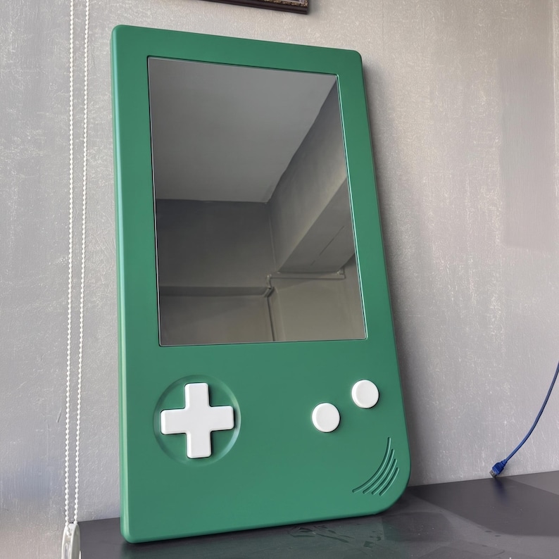 May include: A green mirror shaped like a handheld video game console. The mirror has a rectangular reflective surface and features white buttons and a directional pad. The frame is a solid, vibrant green.