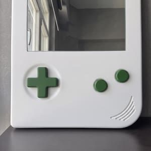 May include: A white mirror in the shape of a retro handheld game console. It features a green directional pad and green buttons. The design includes a curved edge with etched lines, resembling a smile.