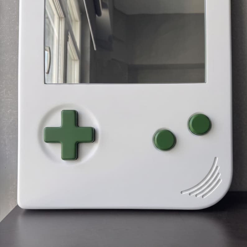May include: A white mirror in the shape of a retro handheld game console. It features a green directional pad and green buttons. The design includes a curved edge with etched lines, resembling a smile.
