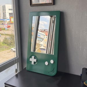 May include: A green mirror in the shape of a classic handheld gaming device. The mirror has a white cross-shaped directional pad and two white buttons. The mirror is rectangular with rounded edges.