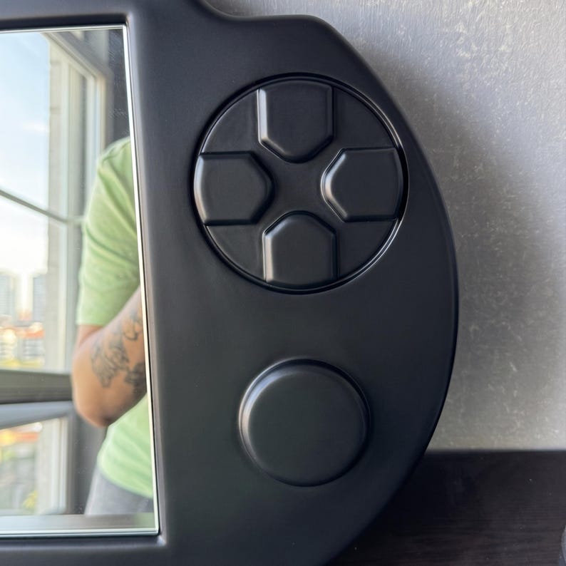 A decorative wall mirror designed in the shape of a black handheld gaming console. The frame includes raised 3D details of a D-pad, action buttons, and joysticks