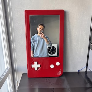 May include: A red, rectangular mirror shaped like a classic handheld video game console. The mirror features a white cross-shaped directional pad and two white circular buttons. The frame is a vibrant red, and the mirror reflects the room.