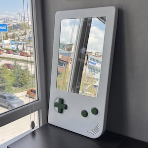 May include: A white mirror shaped like a retro handheld game console. It features a green directional pad and buttons, with a curved speaker design at the bottom. The mirror reflects a cityscape.