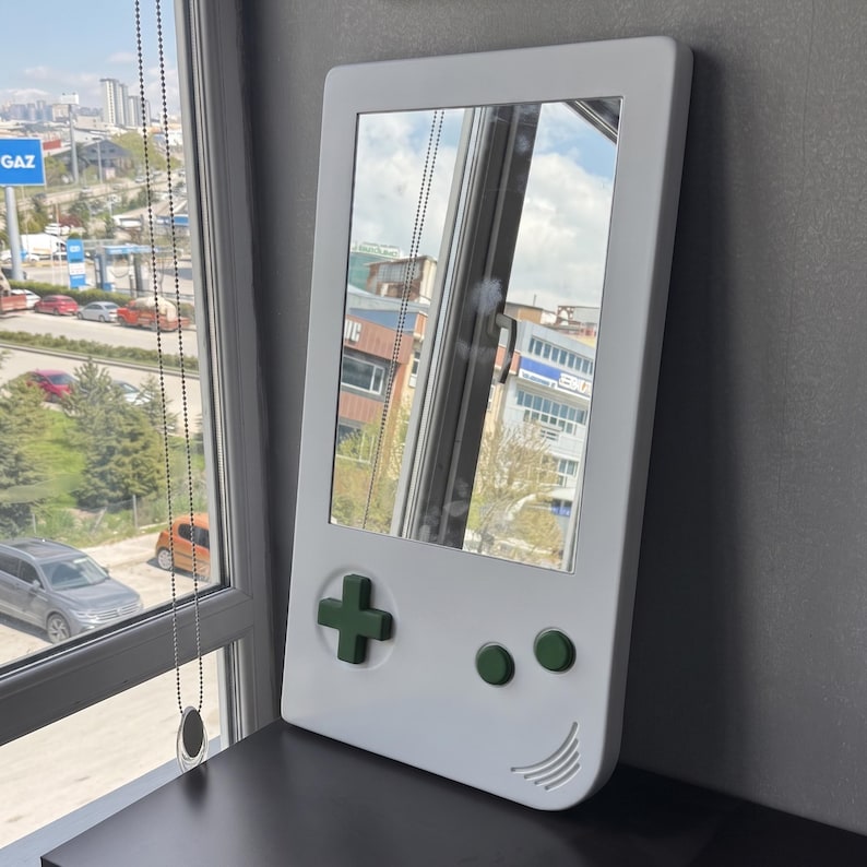 May include: A white mirror shaped like a retro handheld game console. It features a green directional pad and buttons, with a curved speaker design at the bottom. The mirror reflects a cityscape.