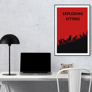 May include: A framed poster with the text "EXPLODING KITTENS" in black letters against a red background. Below the text, black silhouettes of cats are arranged on a dark hill. The poster is displayed on a white desk with a black lamp and laptop.