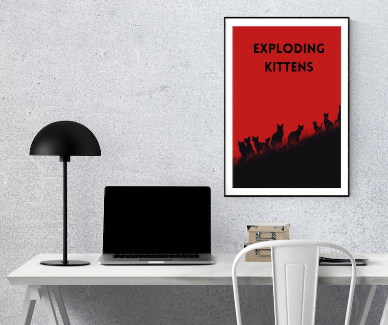 May include: A framed poster with the text "EXPLODING KITTENS" in black letters against a red background. Below the text, black silhouettes of cats are arranged on a dark hill. The poster is displayed on a white desk with a black lamp and laptop.