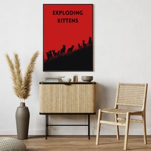 May include: A framed art print featuring the text "EXPLODING KITTENS" above a silhouette of cats on a hill against a red background. The print is displayed above a wooden cabinet and next to a woven chair.
