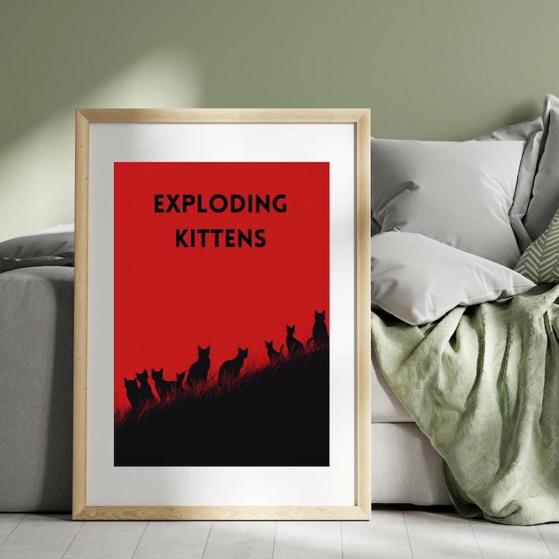 May include: Framed art print featuring the words "EXPLODING KITTENS" in black on a red background. Below the text, a black silhouette of kittens is depicted on a black hill. The frame is a light wood color.