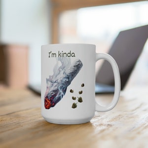 May include: White ceramic mug with the text "I'm kinda" and a graphic of a lit joint with smoke and several buds. The mug has a large handle and is sitting on a wooden surface.