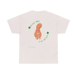 May include: A light beige t-shirt with a graphic of a cat's rear end and the text "Kiss Me I'm Irish" in green. The cat is orange and facing away. Two green shamrocks flank the text.