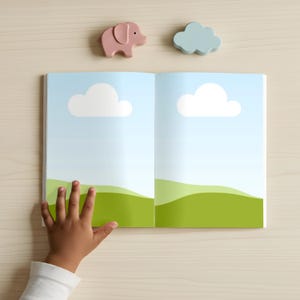 May include: An open book with a blue sky and green landscape illustration, featuring white cloud shapes. A pink elephant and a blue cloud-shaped wooden toy are above the book. A child's hand is touching the book.