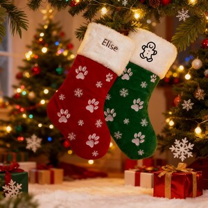 Könnte beinhalten: Zwei Weihnachtsstrümpfe, einer rot und einer grün, hängen an einem Ast. Der rote Strumpf hat weiße Pfotenabdrücke und Schneeflocken sowie den Namen "Elise" auf der Manschette. Der grüne Strumpf hat Pfotenabdrücke und einen Lebkuchenmann.