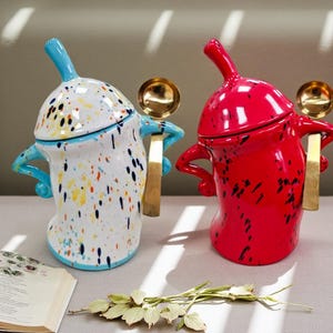 May include: Two ceramic spice jars. One is white with colourful paint splatters, the other is red with black splatters. Each jar has a gold spoon and a blue or red handle. The jars are decorative.