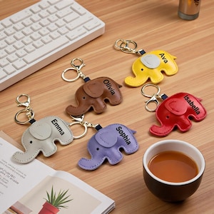May include: A collection of personalized elephant-shaped keychains in various colors, including yellow, brown, red, purple, and cream. Each keychain has a name printed on it, such as "Ava", "Olivia", "Isabella", "Sophia", and "Emma".